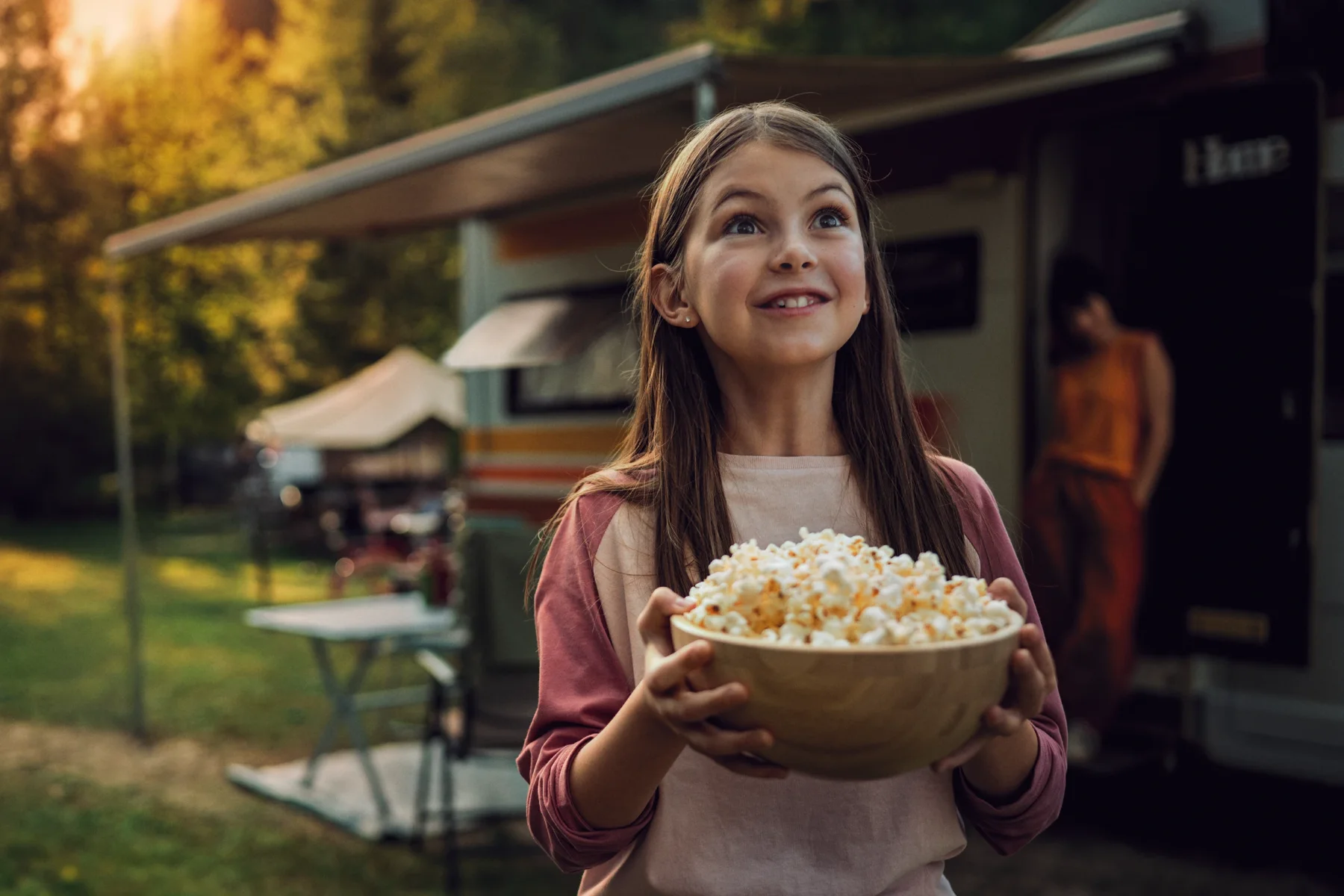 a girl carries a bowl of popcorn in front of her families' caravan